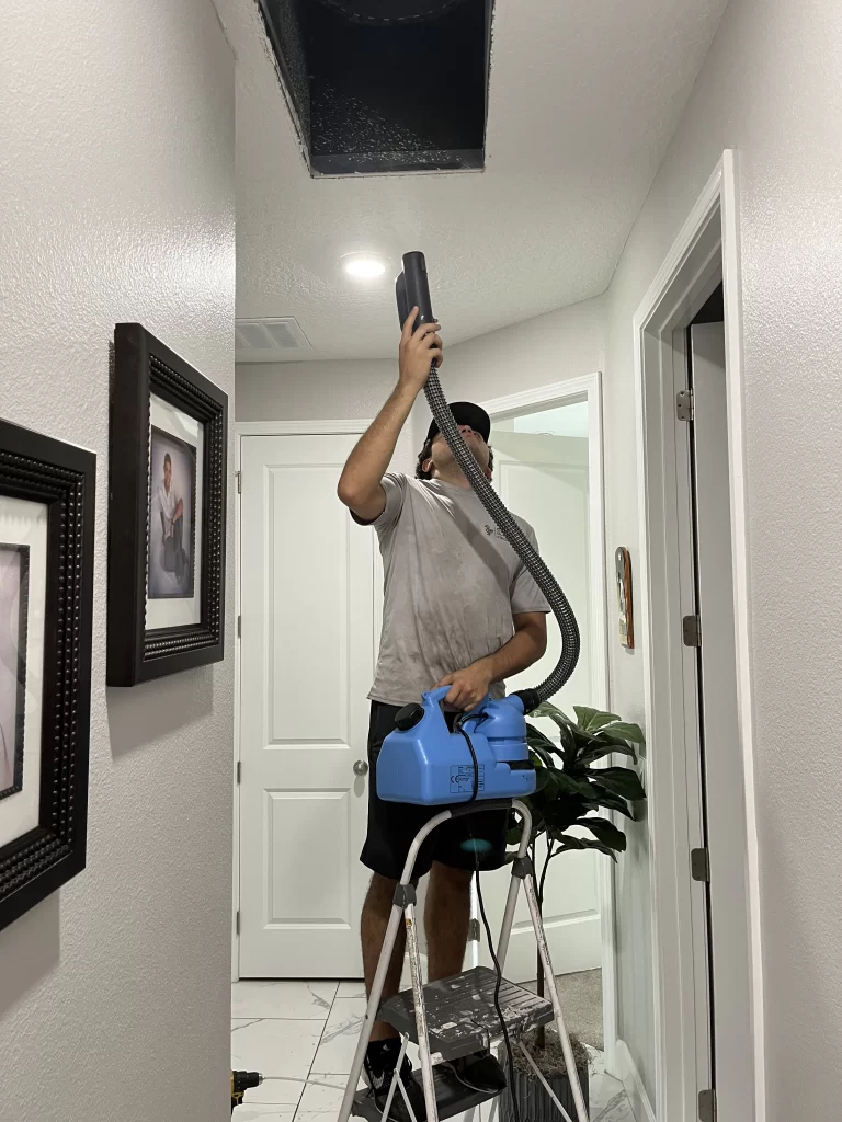Worker holding an air duct sanitizing machine and pointing a hose toward the ceiling at a vent opening.